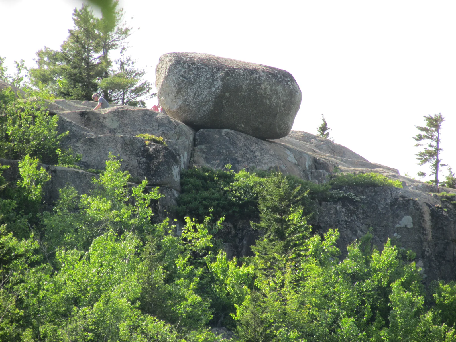 The Bubble Rock boulder in Acadia National Park on the edge of South Bubble, with people beside it, above a wooded rock slope
