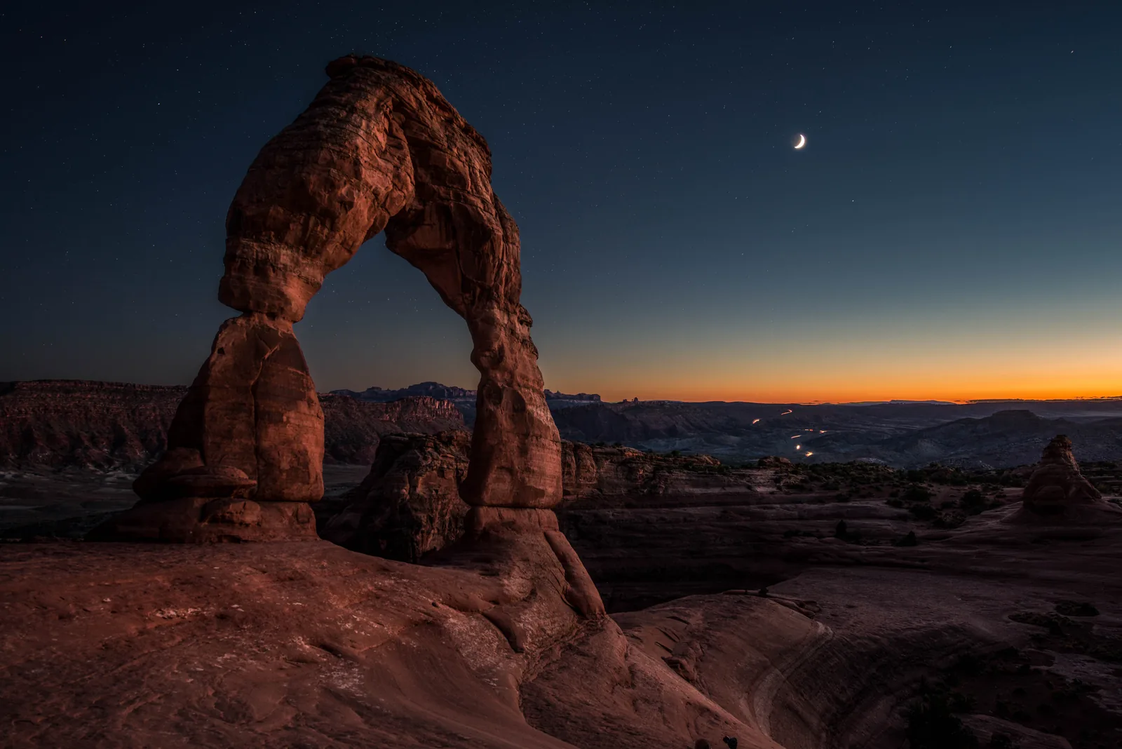 Delicate Arch in Arches National Park at dusk against a deep blue sky with a thin crescent moon