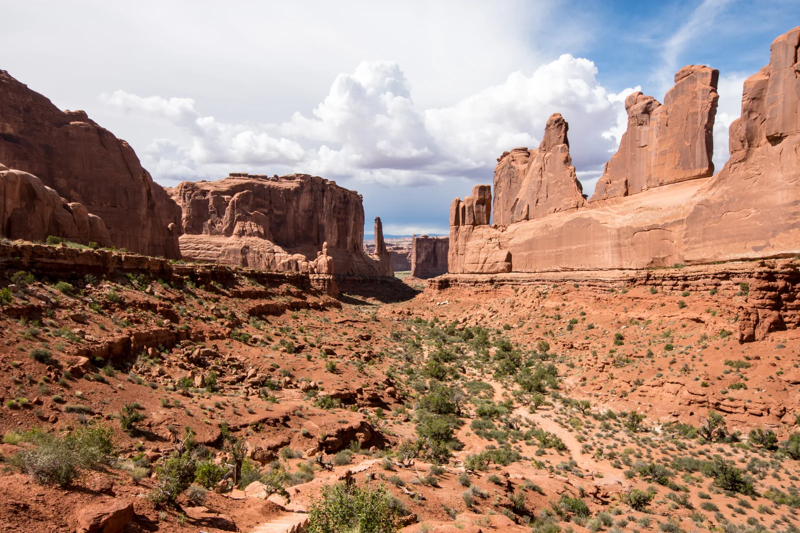 Sheer red sandstone walls in Arches National Park on both sides of a dry valley under cumulus clouds
