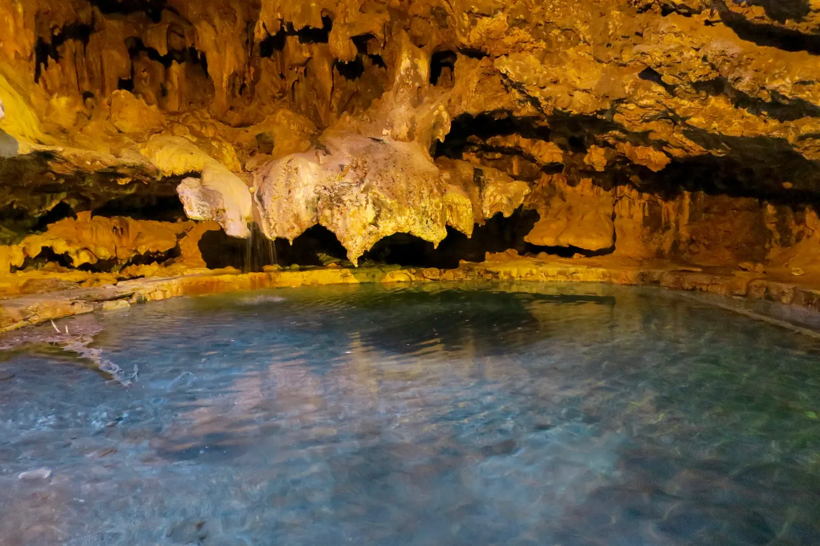 Clear thermal water in the Cave and Basin cave in Banff National Park below golden-brown lit rock formations.
