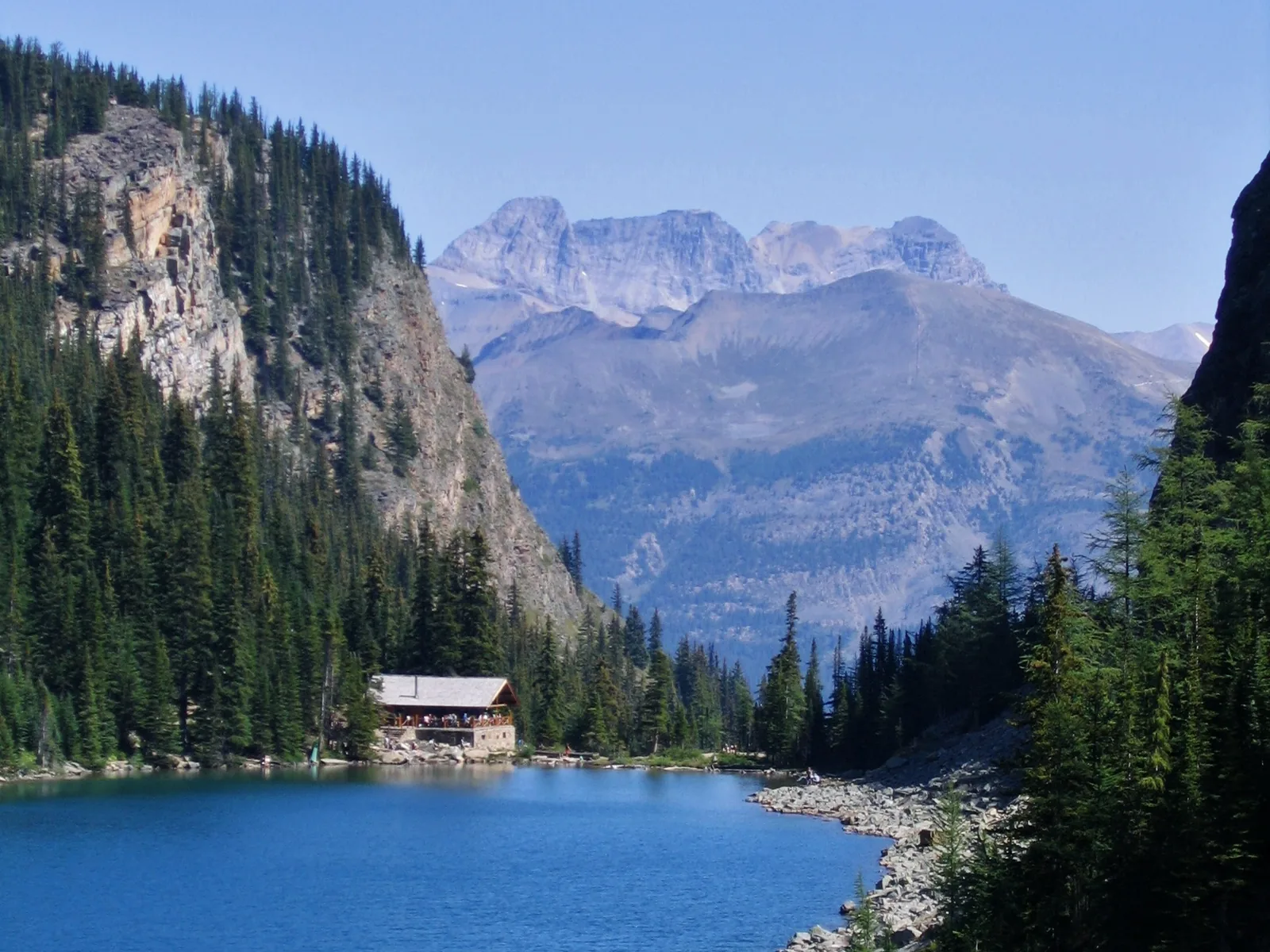 The teahouse at Lake Agnes in Banff National Park on the shore of a dark blue mountain lake below a rock face.