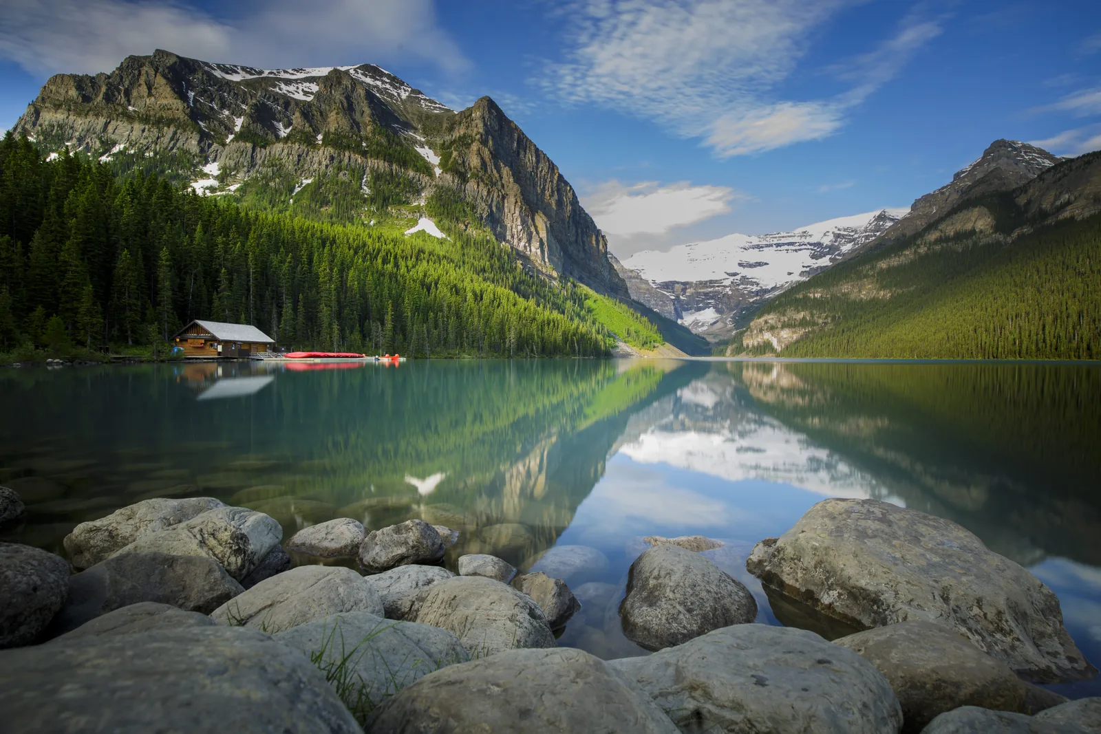 Lake Louise in Banff National Park with the boathouse and red canoes, the Victoria Glacier behind, shore rocks in the foreground.