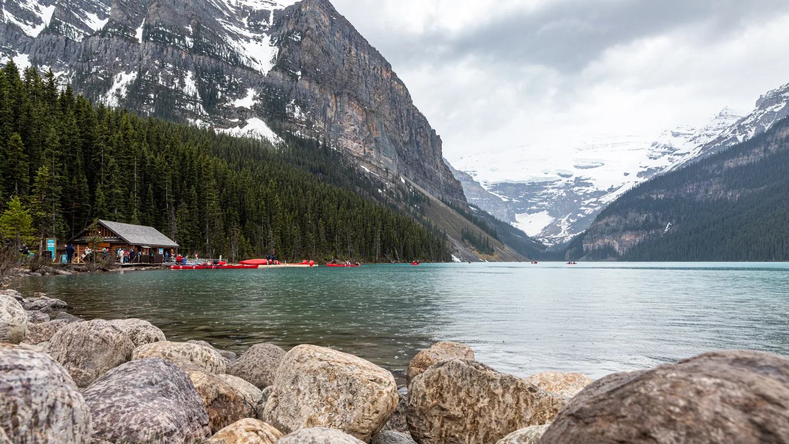 Lake Louise in Banff National Park from the east shore: boulders in the foreground, boathouse and canoes on the shore, the glacier behind.