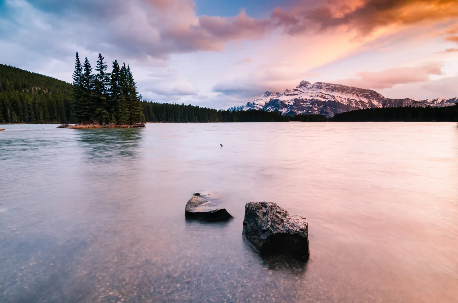 Two Jack Lake in Banff National Park in evening light: pink, glassy water with two stones, a forested island and snowy peaks.