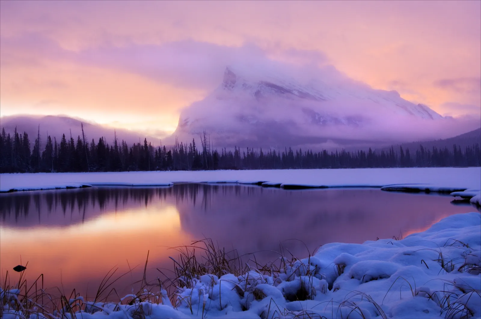 Snowy shore of the Vermilion Lakes in Banff National Park at sunrise, a cloud-covered mountain reflected in pink and violet in the water.