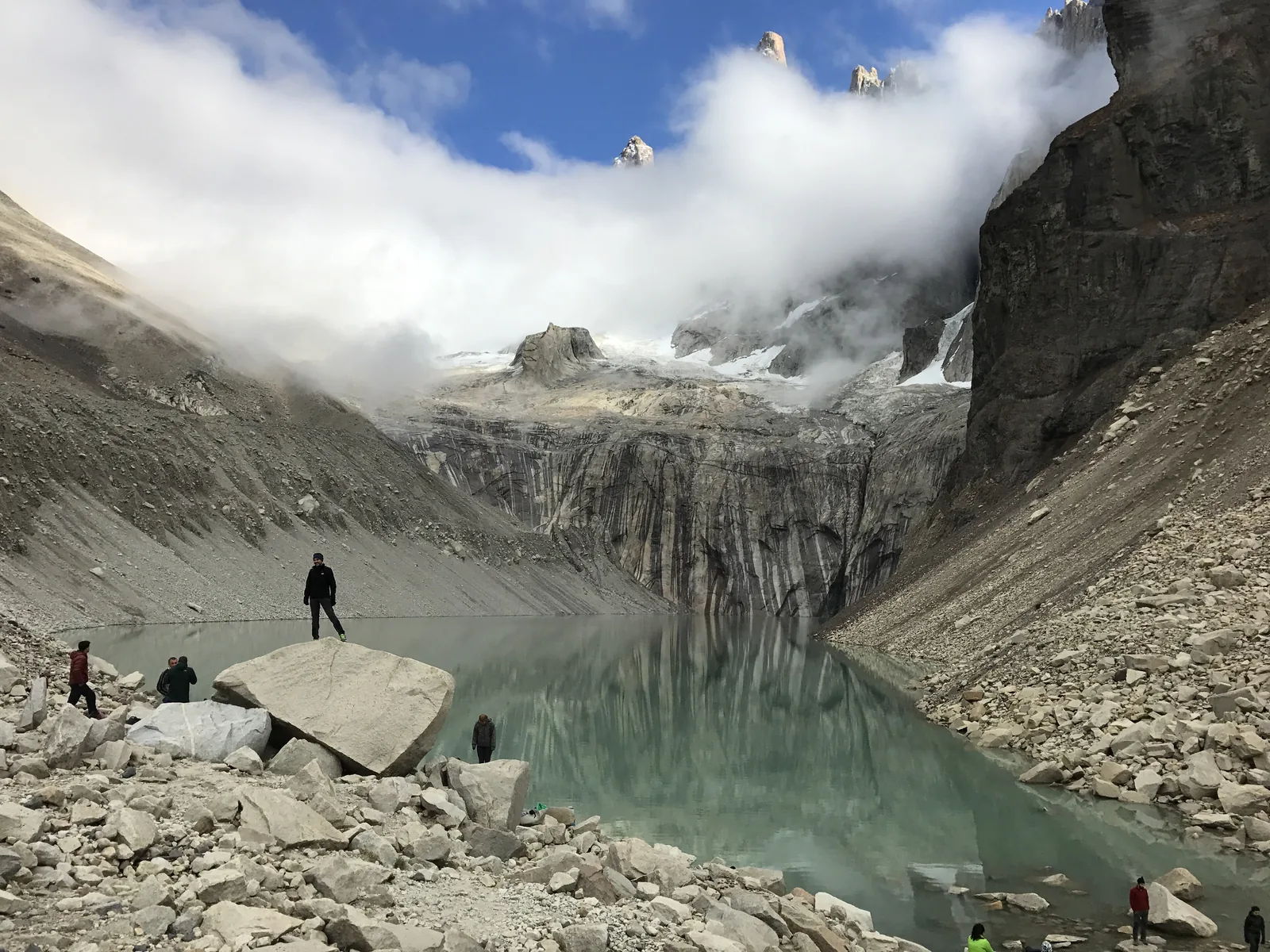 Hikers at the Mirador Base Torres in Torres del Paine National Park: boulders by the green glacial lake, clouds on the granite towers above
