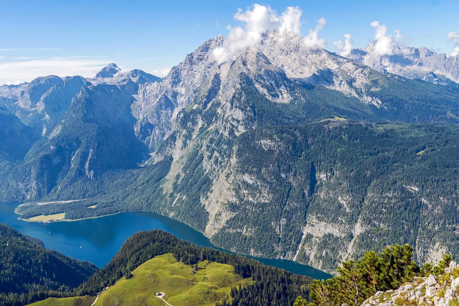 View down from the Jenner onto the dark blue Lake Königssee, above it a massive limestone massif with clouds on the summit