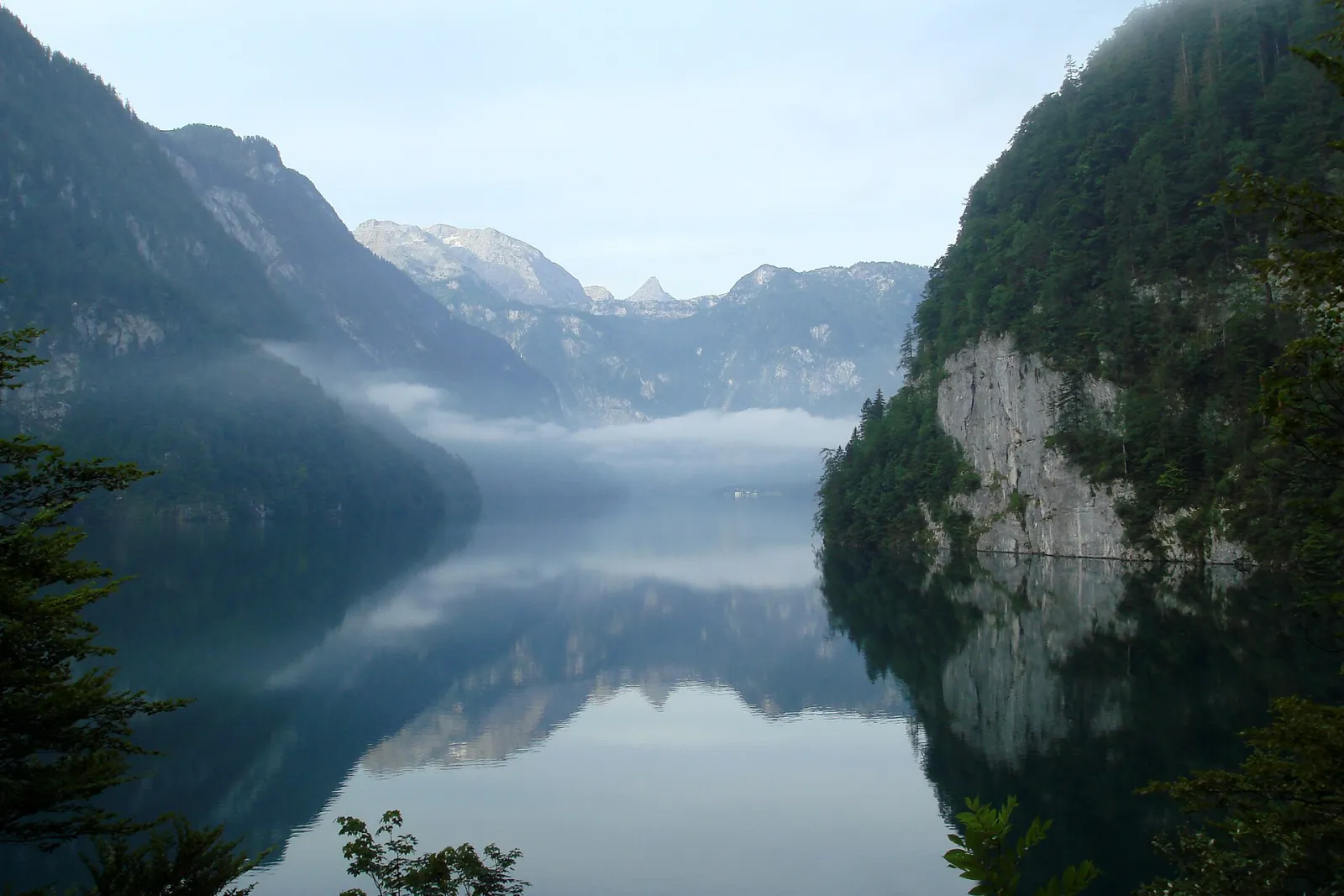 Lake Königssee in Berchtesgaden National Park shortly after sunrise from the Malerwinkel viewpoint: still water reflects the steep, wooded shore walls, with banks of mist and bright summits behind