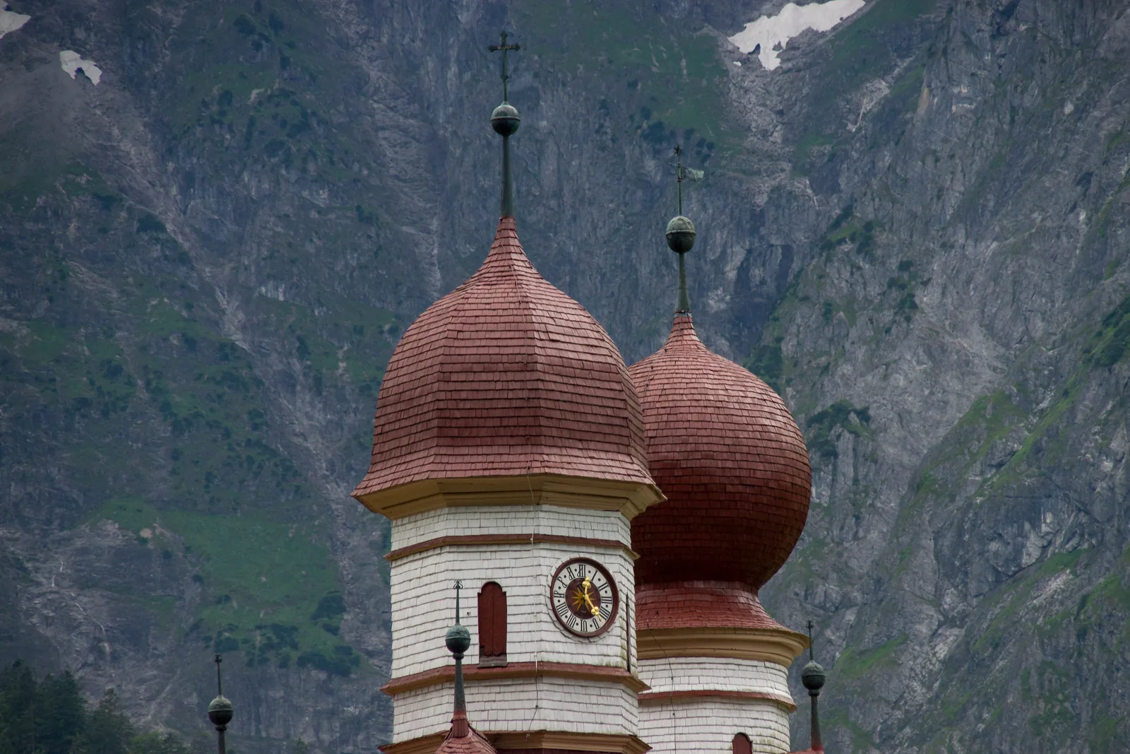 The two onion domes of the church of St. Bartholomä in Berchtesgaden National Park, with red shingles and a golden clock in front of a grey rock face