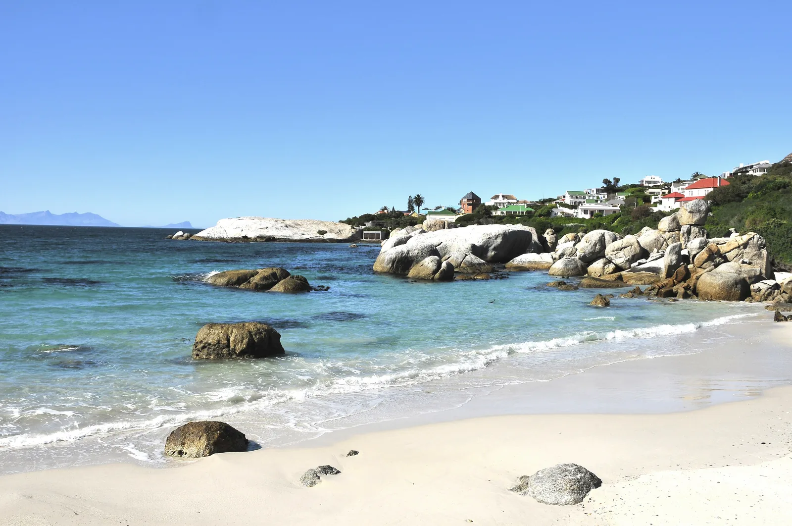 Foxy Beach at Boulders Beach with white sand, round granite boulders and clear water, houses of Simon’s Town behind