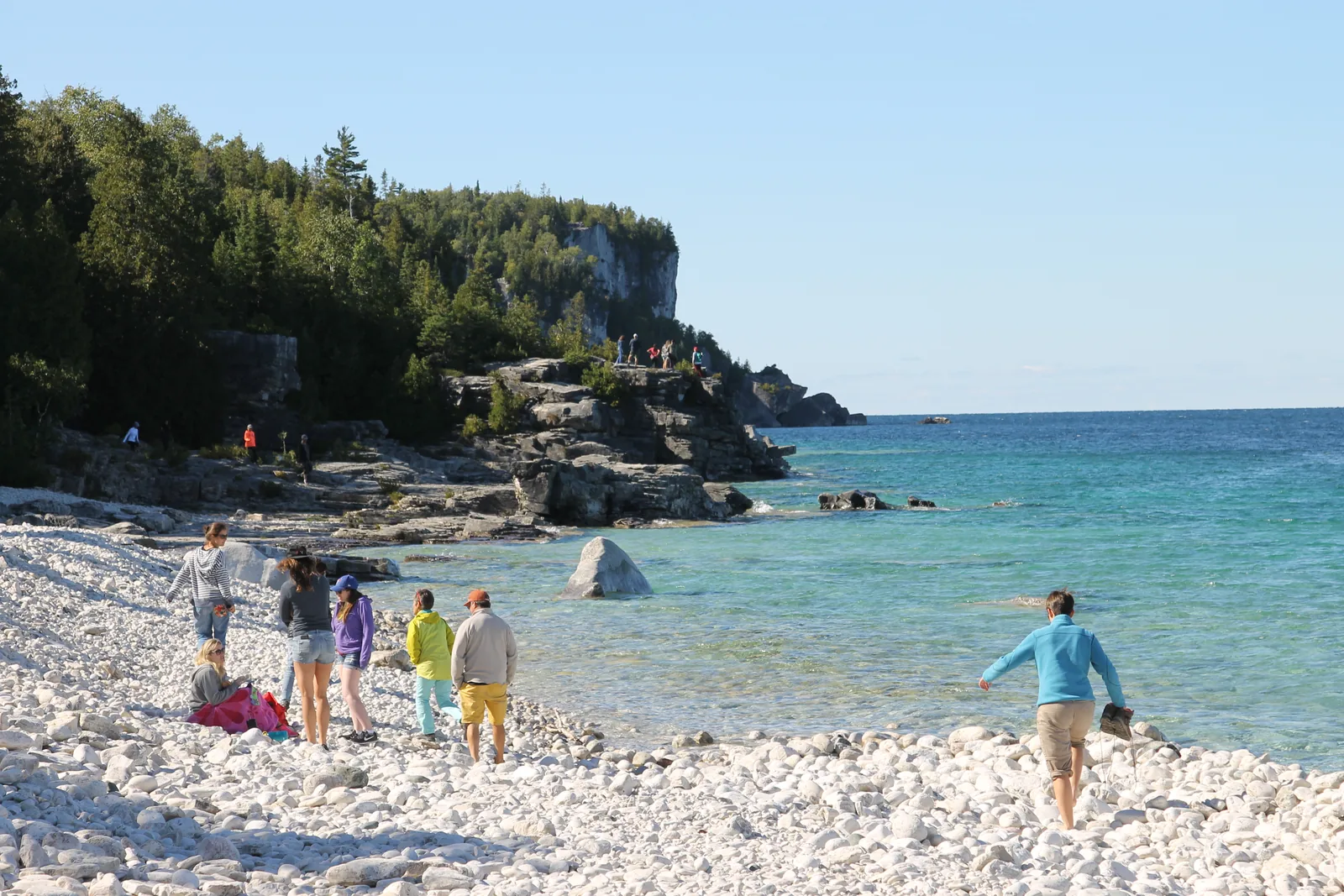Pale cobble beach in Bruce Peninsula National Park on a turquoise bay, wooded limestone cliffs behind, a few people at the water