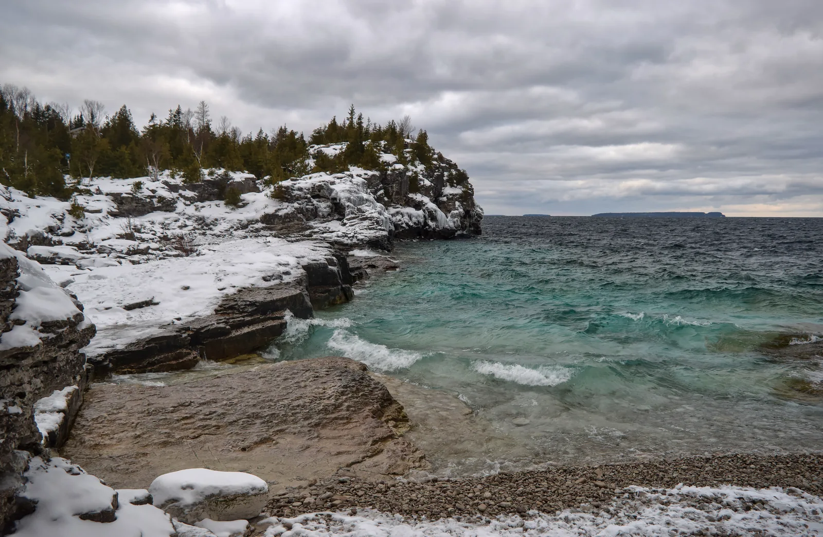 Snow-covered limestone terraces at Indian Head Cove in Bruce Peninsula National Park, turquoise water below, a wooded headland behind under a grey sky