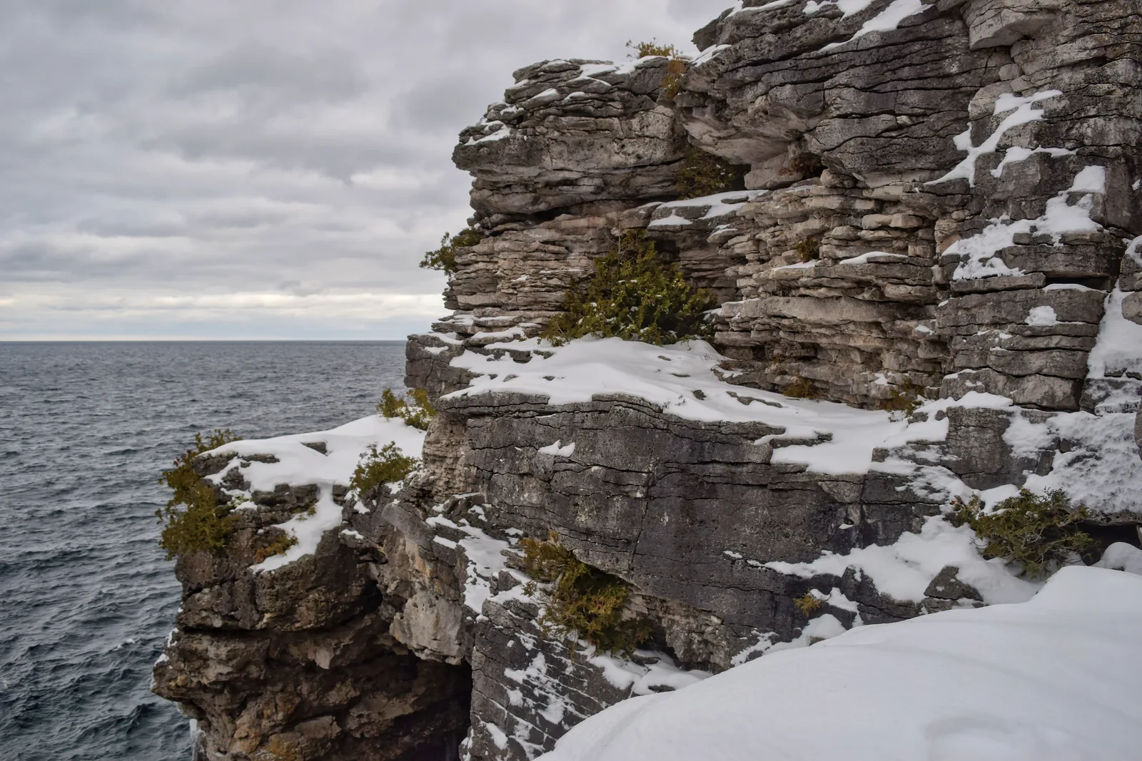 Snow-covered, horizontally layered limestone cliffs drop into dark Georgian Bay in Bruce Peninsula National Park, twisted cedars growing in the rock crevices