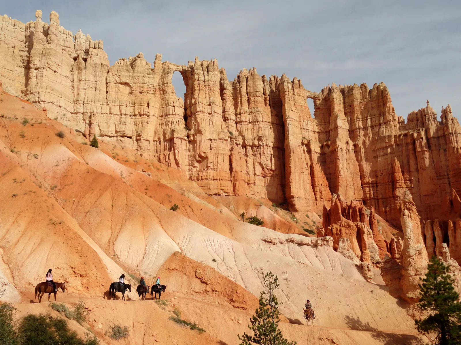 A group of riders on the Peekaboo Loop in Bryce Canyon National Park below a tall rock wall pierced by windows, with pale, furrowed slopes in front