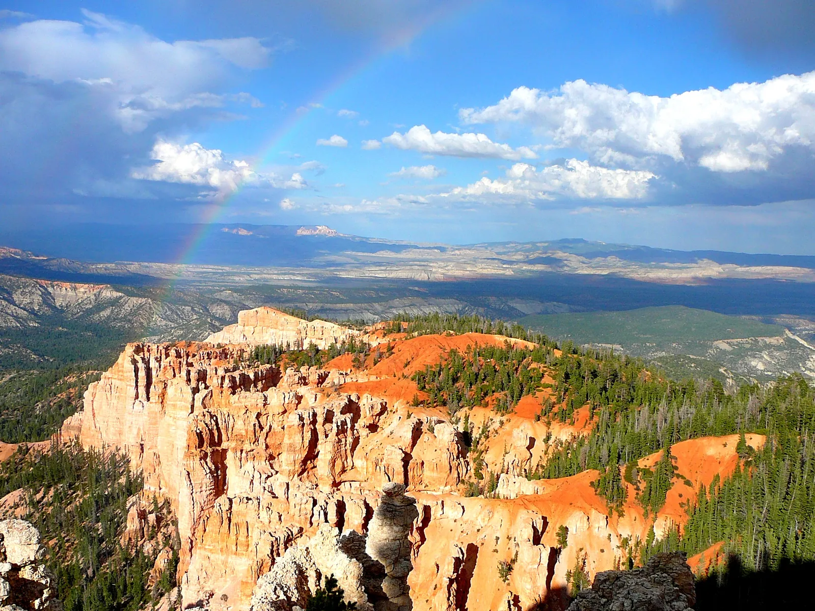 A rainbow over a wide landscape in Bryce Canyon National Park, with pale cliffs, red slopes and dark conifer forest on the plateau edge below