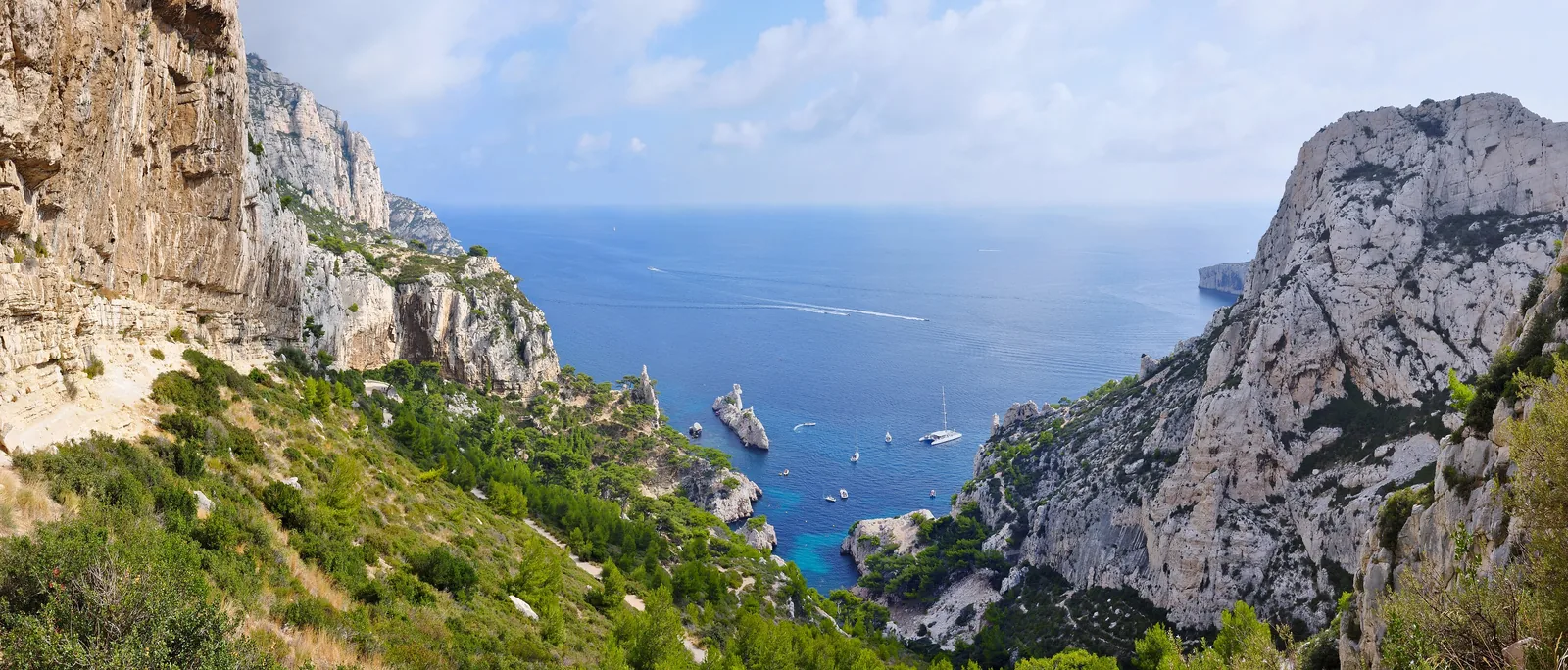 Panorama into the Calanque de Sugiton in Calanques National Park from the hiking path: limestone rocks, green slopes and boats in blue water
