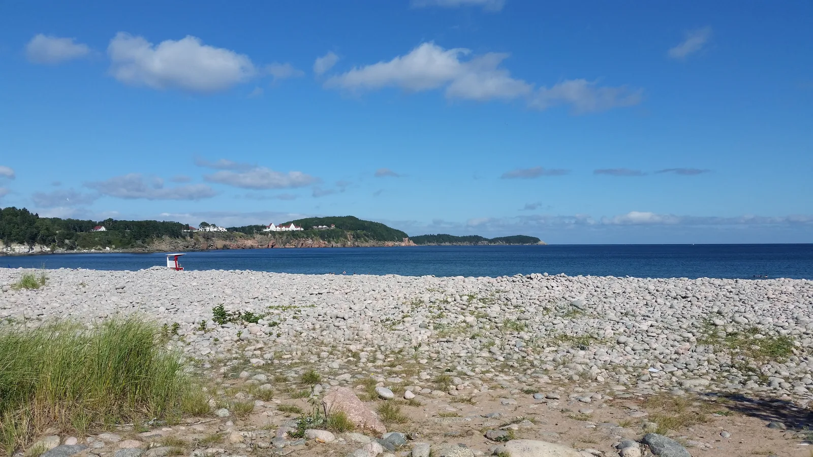 Wide pebble beach at Freshwater Lake in Cape Breton Highlands National Park under a blue sky, with a low forested headland and the open sea behind