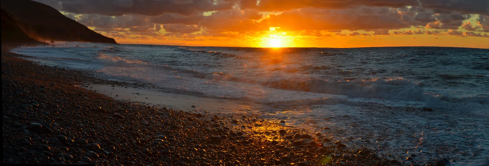 Sunset over the water seen from a dark pebble beach; a cliff coast runs into the picture on the left