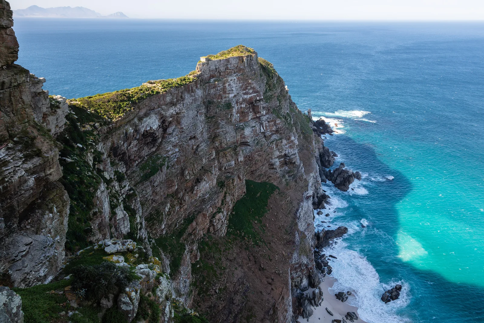 Steep, green cliffs at the Cape of Good Hope drop into turquoise sea with white surf at Cape Point