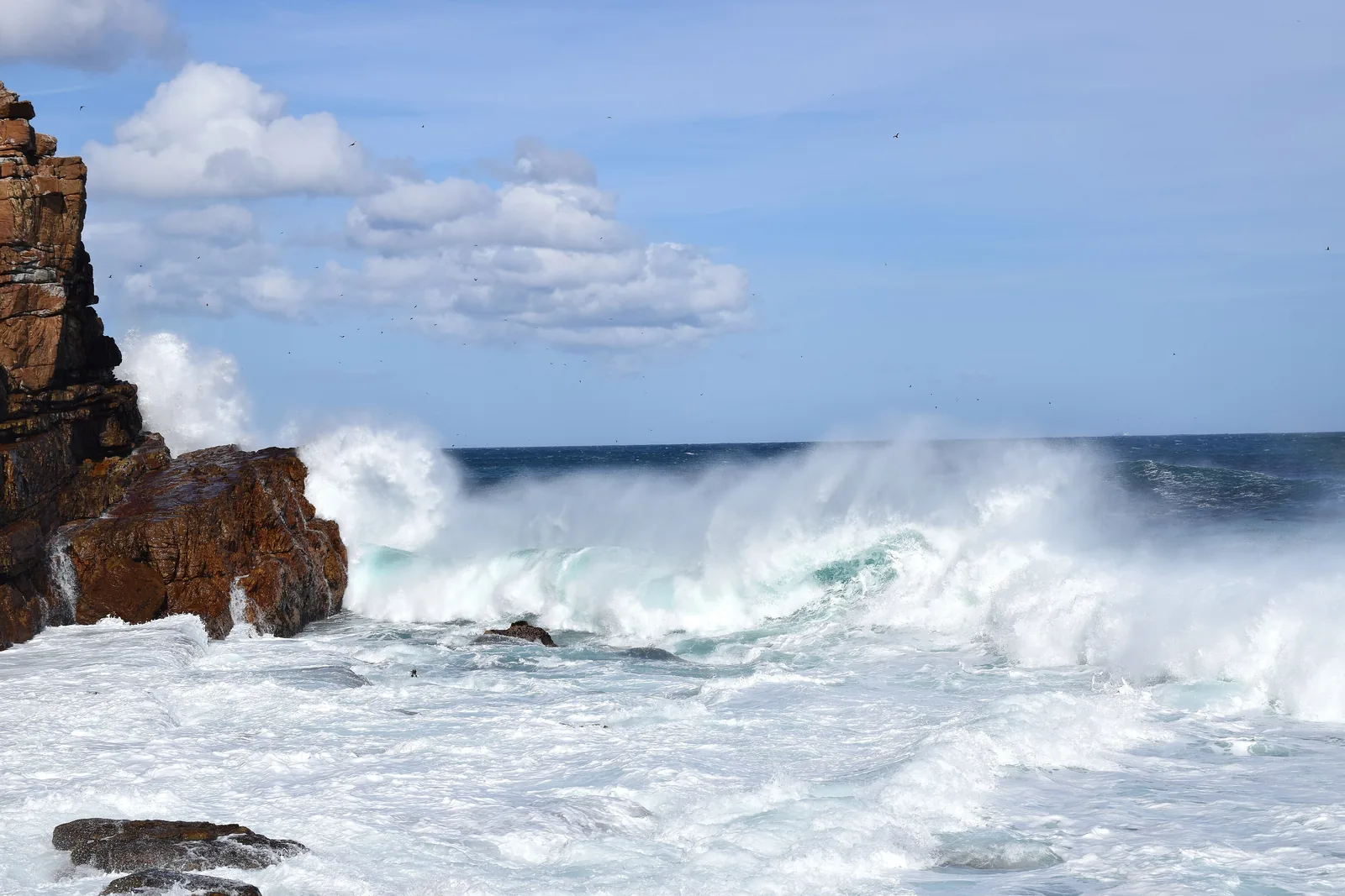 High waves break on dark rocks at the Cape of Good Hope, spray rising against a blue sky