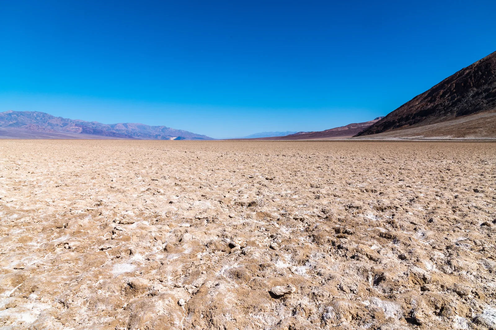 Broken white and brown salt crust of Badwater Basin in Death Valley National Park under a deep blue sky, with a dark mountainside on the right
