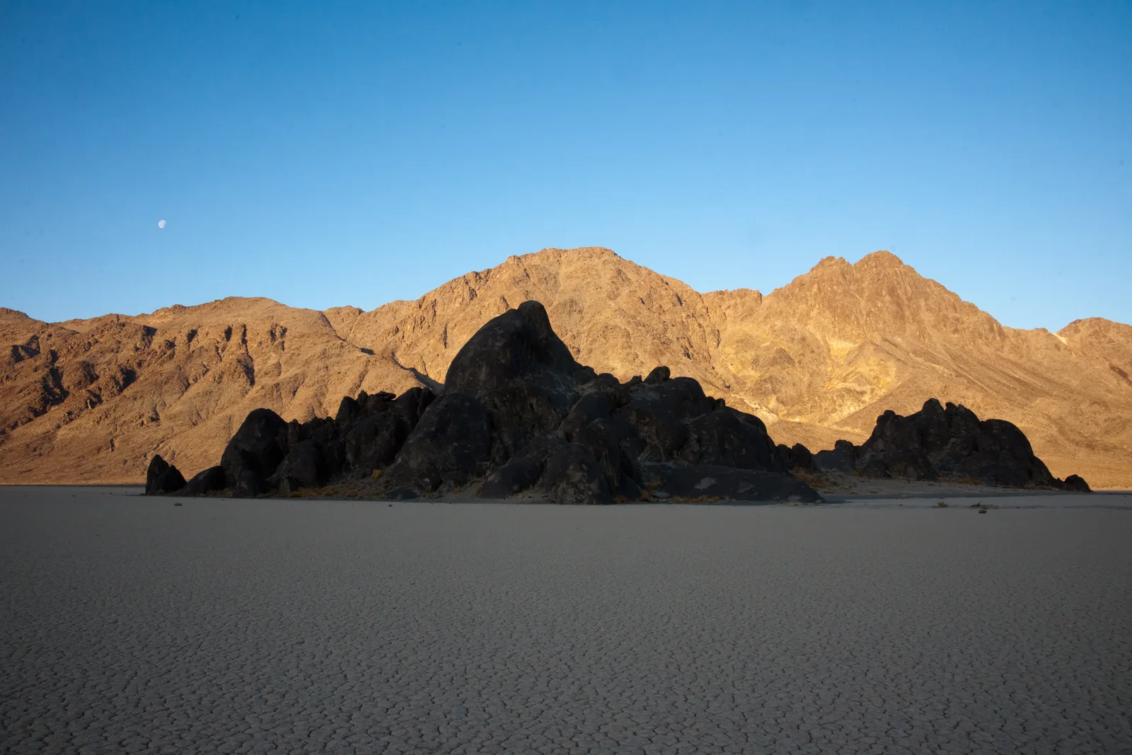 The dark rock island of The Grandstand rises from the flat grey Racetrack Playa in Death Valley National Park, with mountains lit by the morning sun behind