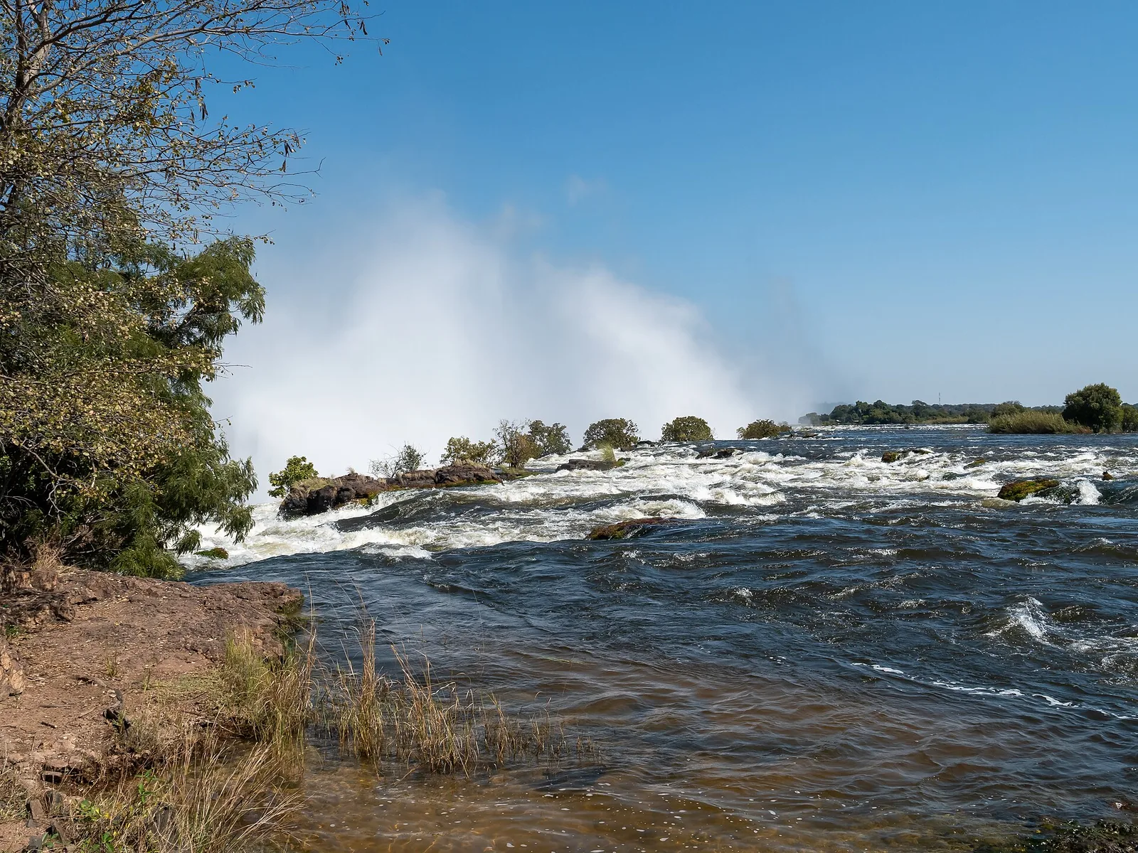 The Zambezi flowing over rocks on the Zambian side towards the edge of Victoria Falls, with spray rising behind