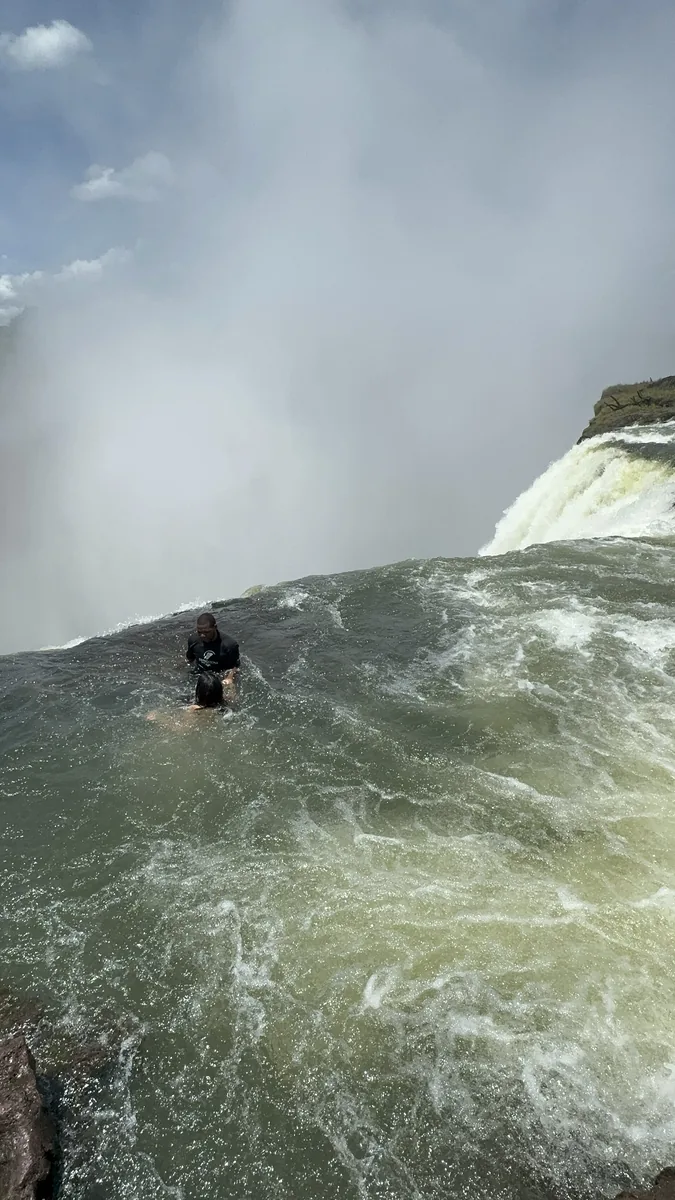 A swimmer in Devil's Pool on the edge of Victoria Falls, the green water of the pool in front and the spray behind