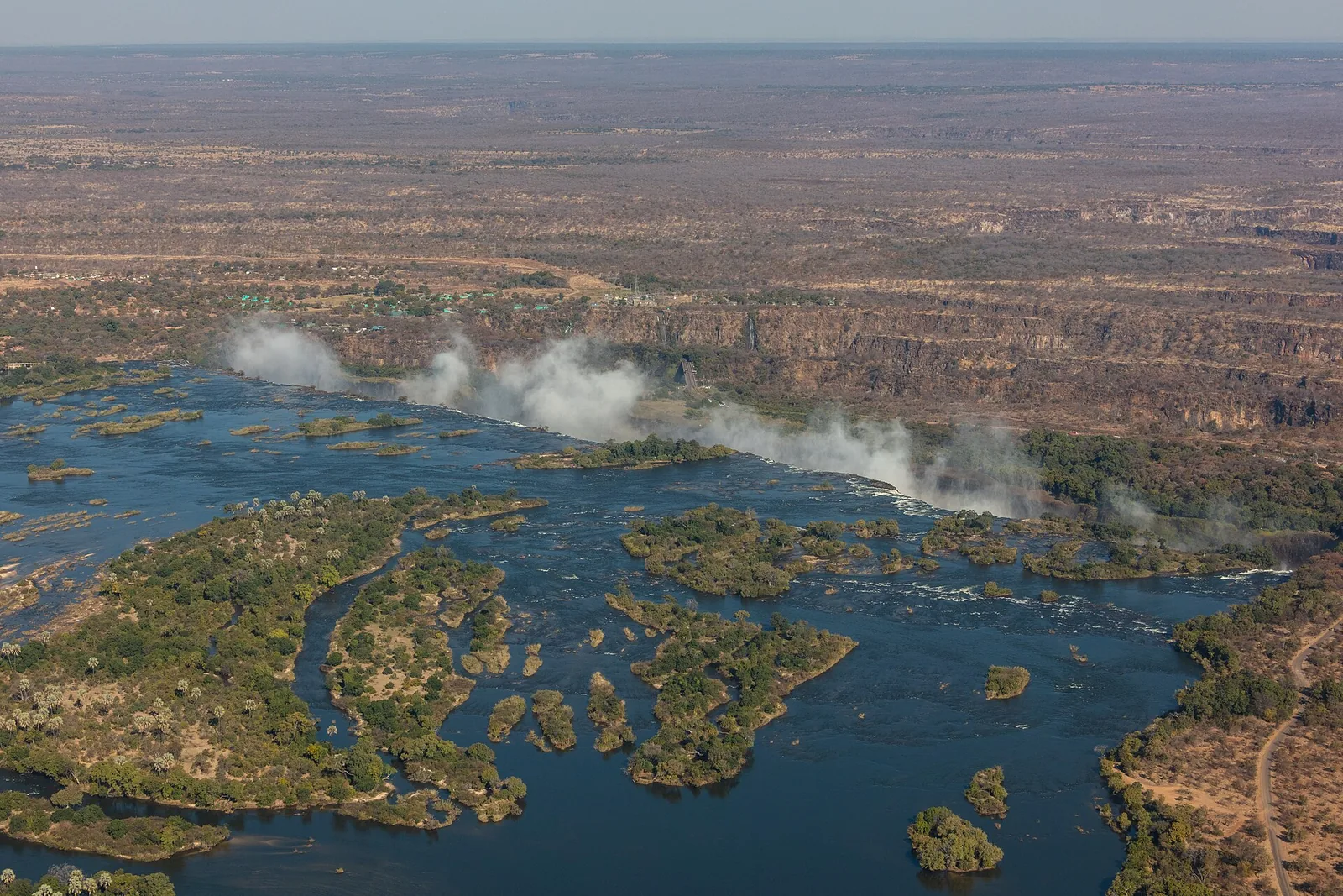 Aerial view of the Zambezi above Victoria Falls with wooded islands reaching to the edge, clouds of spray in the background