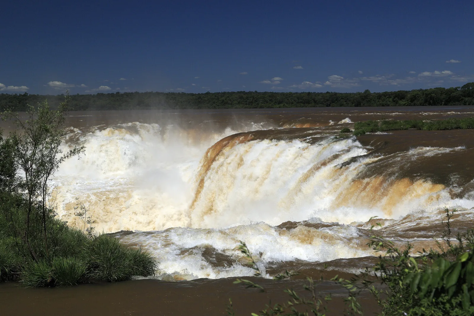 The Iguazú plunges with brown floodwater into the Devil's Throat, spray rising over the edge, the Argentine bank behind