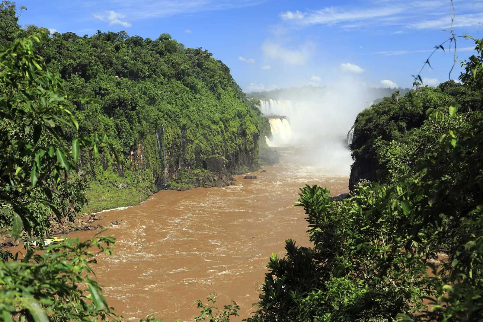 The Iguazú flows brown through a wooded gorge, with spray rising from the Devil's Throat in the background
