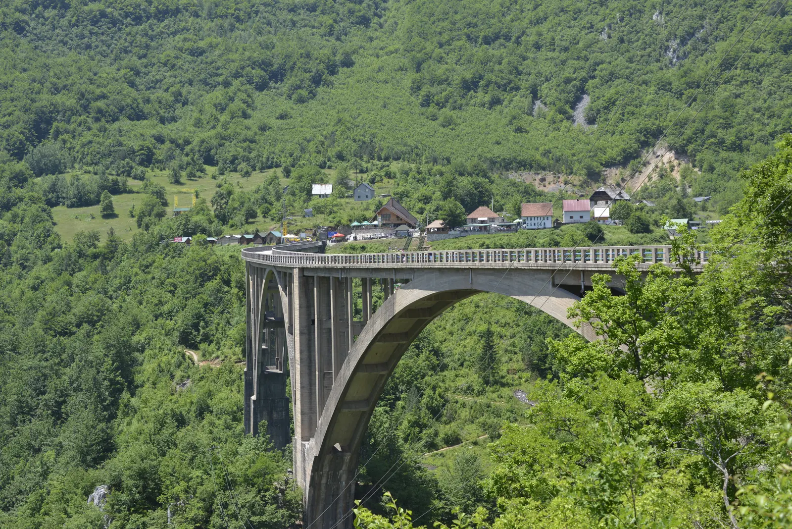 A high concrete arch bridge spans a densely wooded gorge, a few houses at the end of the bridge
