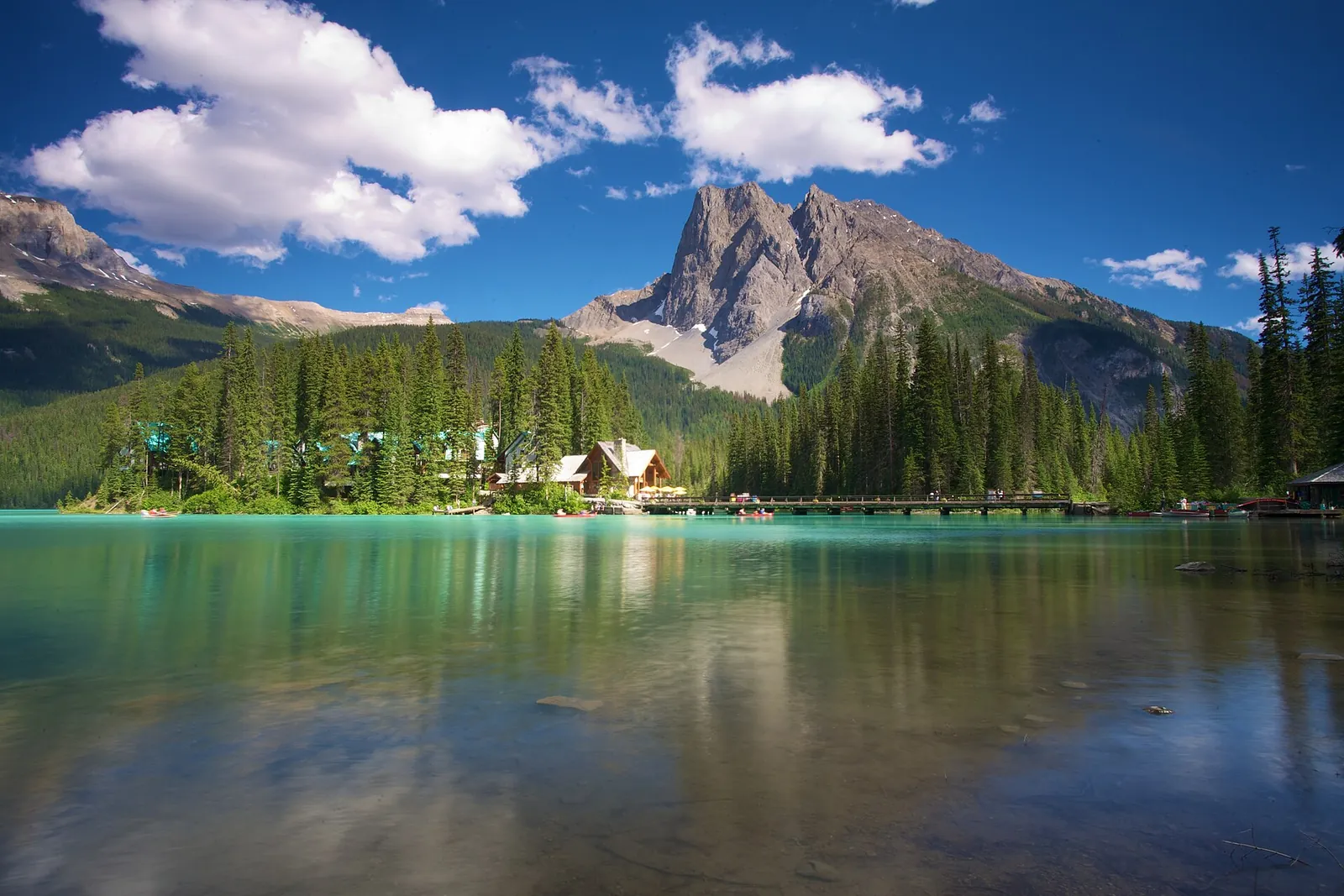 Emerald Lake in Yoho National Park with the lodge on its wooded point below a rocky peak, calm water in the foreground.