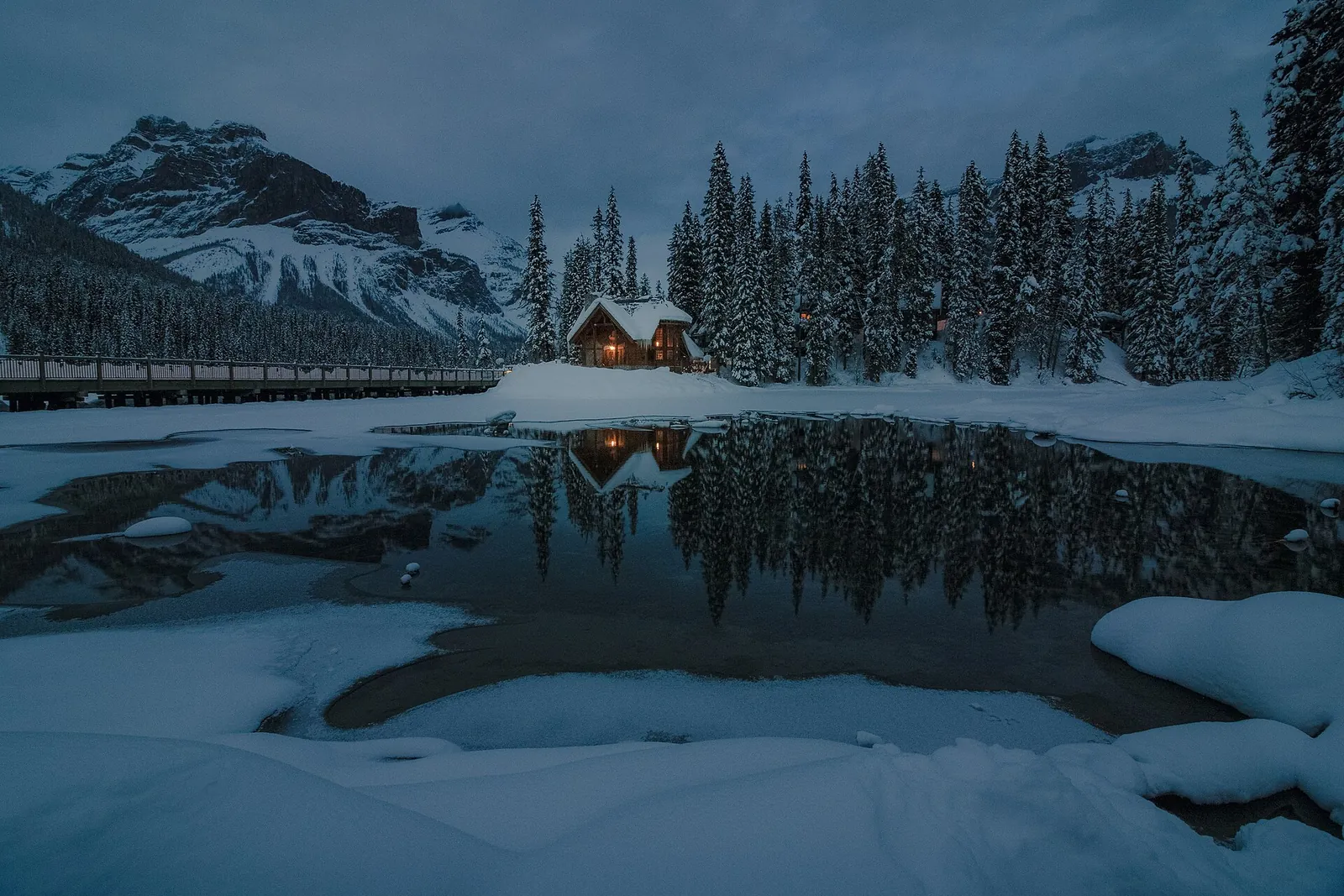 Emerald Lake in Yoho National Park in winter: snowy shore, dark open water and a lit cabin among the firs.