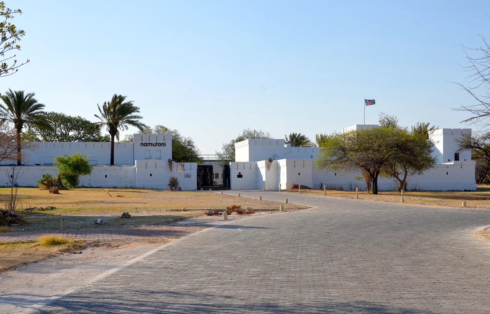 The whitewashed Namutoni Fort in Etosha National Park, with corner towers and battlements behind a palm tree, a well-kept lawn in front.