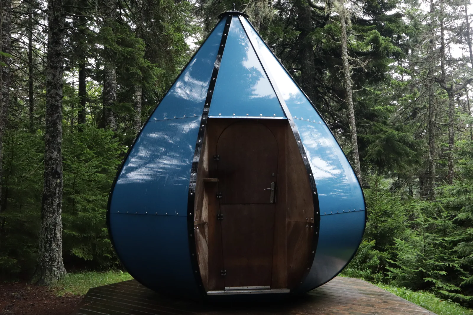 An Ôasis in the forest: a large teardrop-shaped pod with a reflective blue shell and a wooden door, raised on stilts among conifers