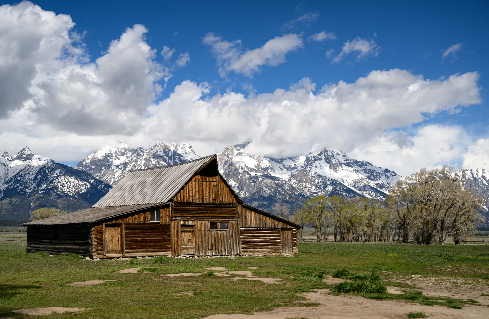 The wooden Moulton Barn in Grand Teton National Park stands in an open meadow, with the snow-covered Teton Range under cumulus clouds behind