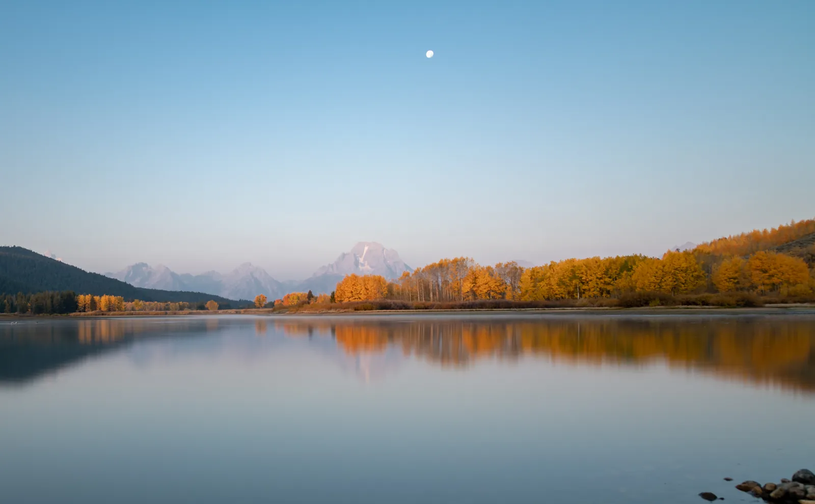 The Snake River at Oxbow Bend in Grand Teton National Park mirrors the morning sky on a still day, with yellow autumn cottonwoods on the bank and the mountain range behind