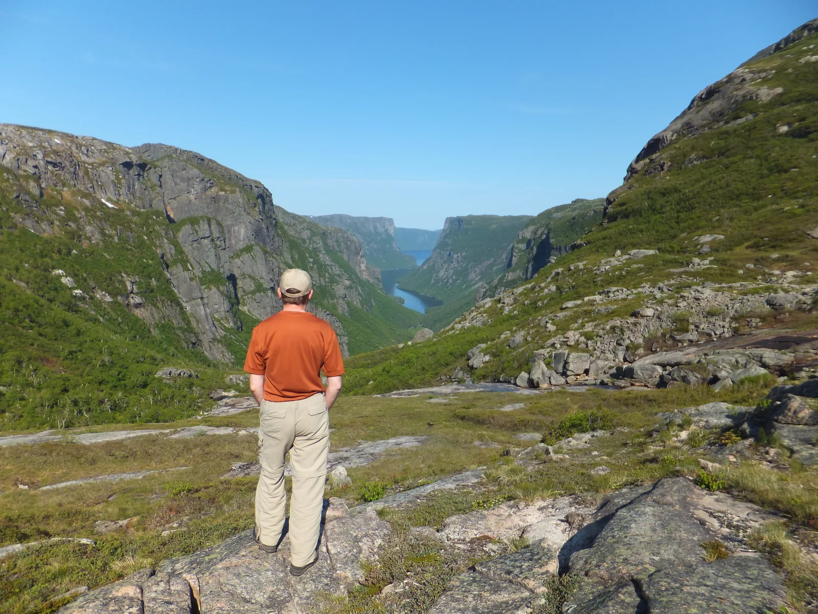 A hiker in an orange jacket looks down from the rocky slope at the end of the fjord in Gros Morne National Park into the long, narrow valley of Western Brook Pond between steep rock walls
