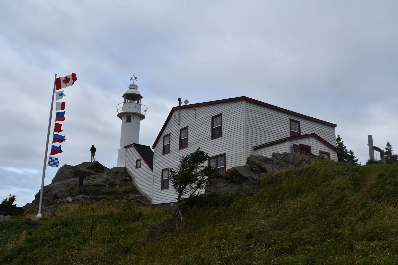 The white Lobster Cove Head lighthouse in Gros Morne National Park with the keeper's house on a rocky headland, in front a flagpole with signal flags