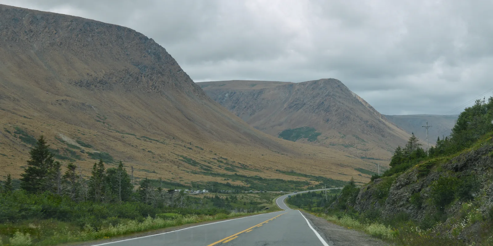 A road leads into a wide valley in Gros Morne National Park; on the left rises the bare, brownish-orange slope of the Tablelands, on the right green conifer forest