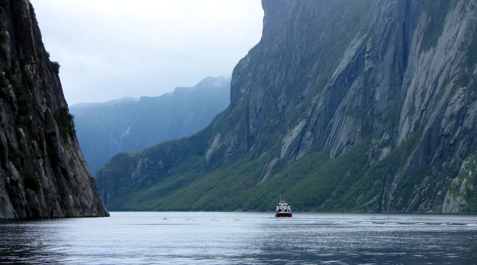 Sheer, dark rock walls frame narrow Western Brook Pond in Gros Morne National Park; in the distance a small tour boat crosses the still water under low clouds