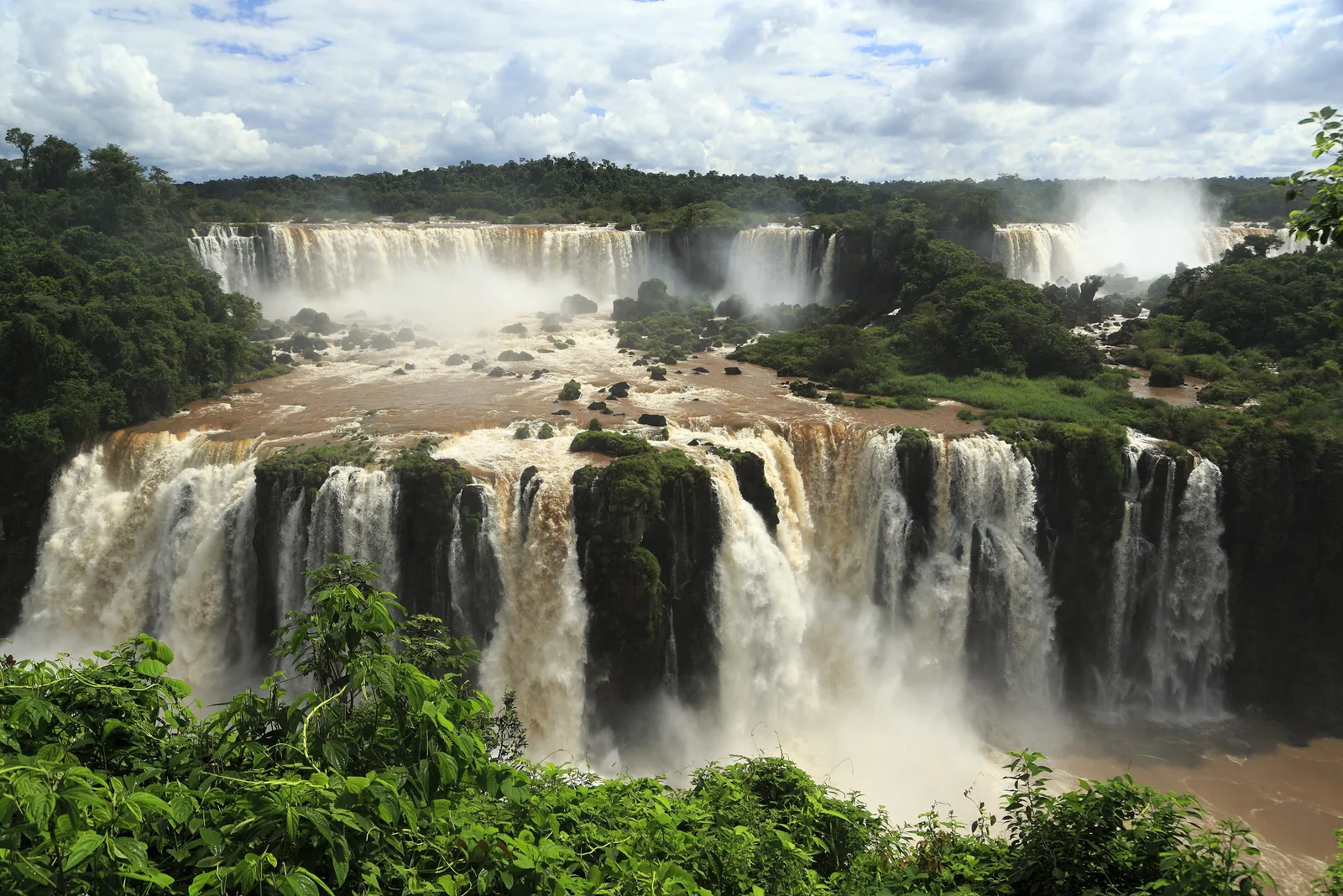 From the Brazilian side of Iguaçu National Park you look over a tier of many white waterfalls to the rocks and forest behind