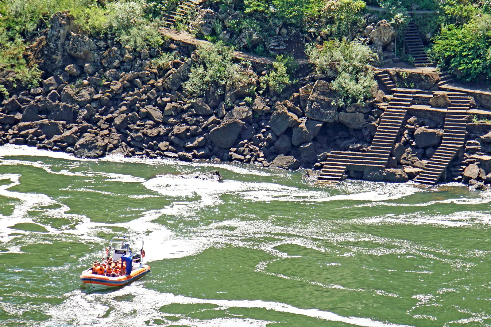 An inflatable boat crosses green water below Iguazu Falls, with steps climbing over rocks on the bank