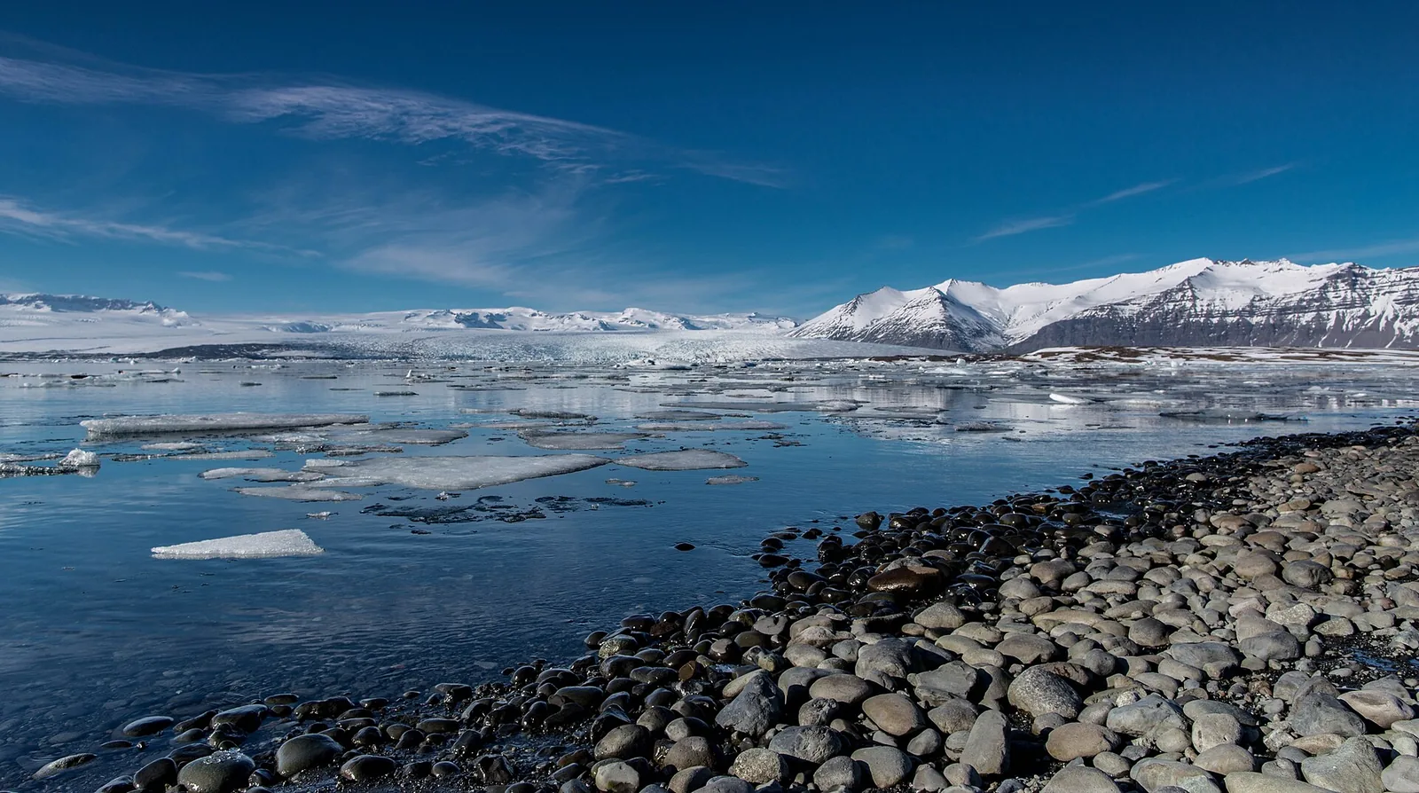 Jökulsárlón glacier lagoon with drifting icebergs in front of Breiðamerkurjökull, a pebble shore in the foreground, snow-covered mountains behind