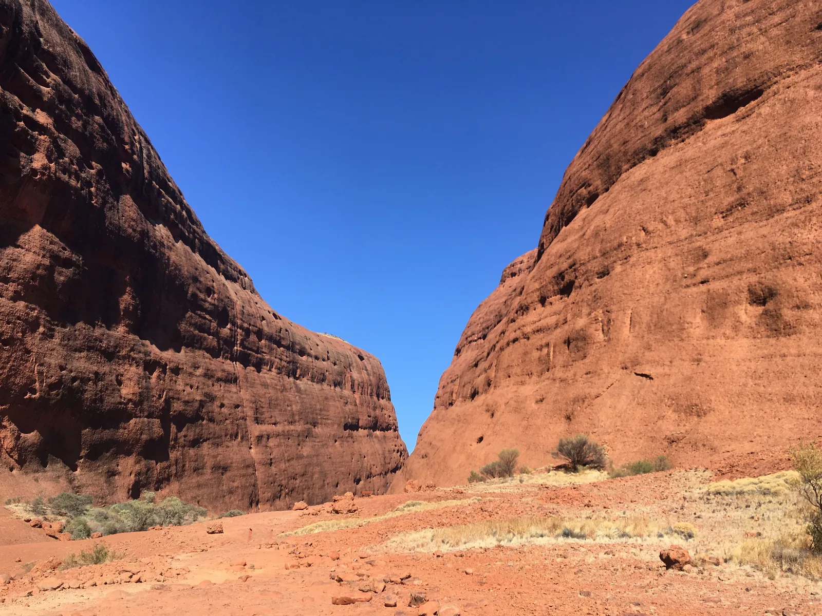 Waḻpa Gorge at Kata Tjuṯa in Uluṟu-Kata Tjuṯa National Park: two high red-brown rock walls to the left and right, a strip of blue sky between them, red sand and grass below