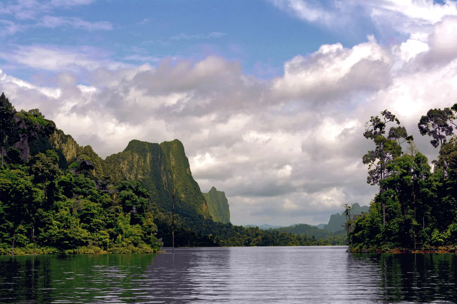A calm arm of the lake between densely wooded shores, a steep limestone tower under white clouds in the background