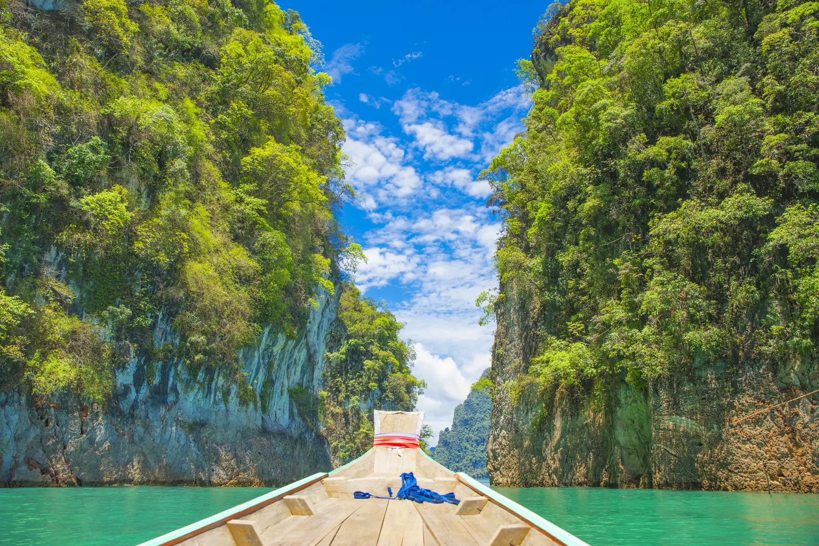 View over the bow of a wooden longtail boat onto turquoise water between two sheer, densely overgrown limestone walls