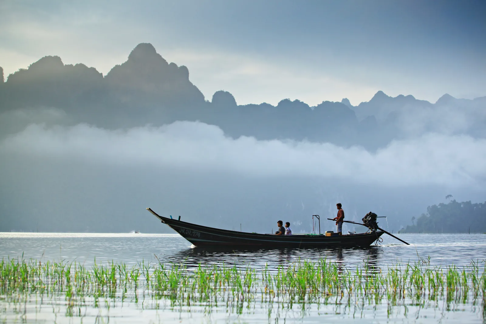 A narrow longtail boat with a standing boatman glides over the still lake, banks of mist in front of jagged limestone mountains behind