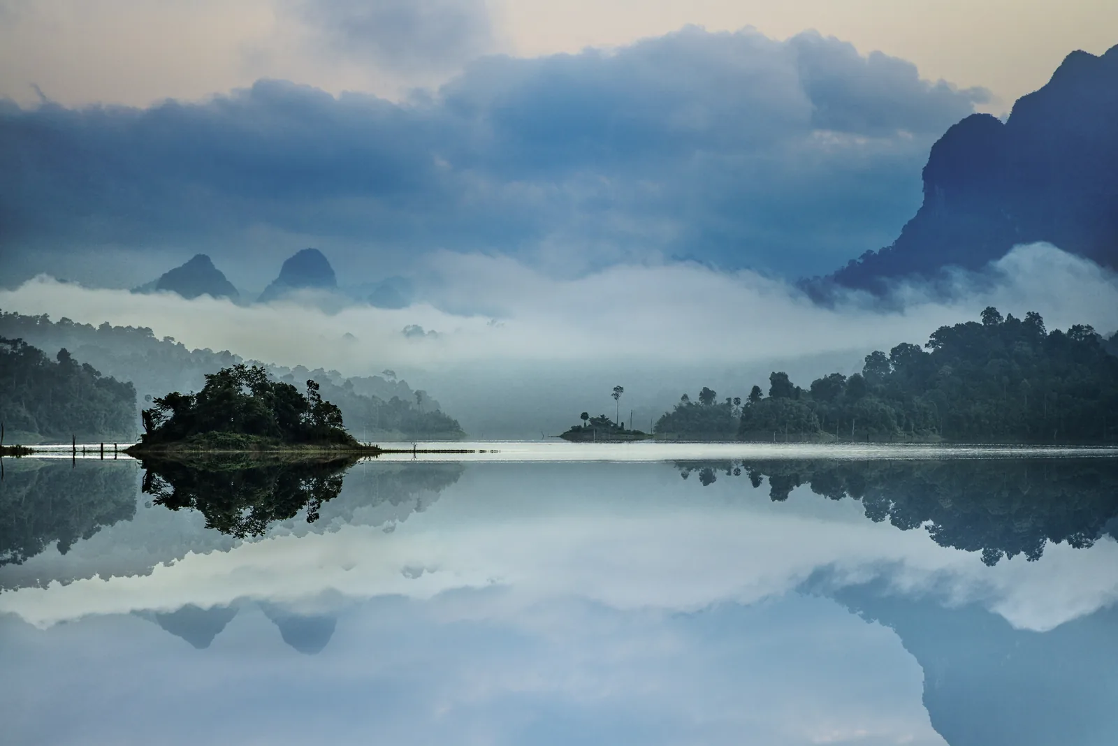 A mirror-smooth lake in morning mist, a small wooded island and behind it the silhouettes of jagged limestone mountains reflected in the water
